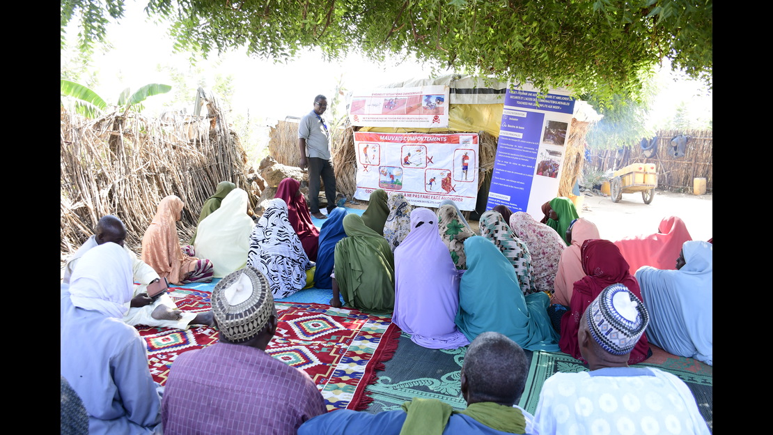  A group of people is sitting on the ground on colorful rugs and mats, arranged in the shade of a large tree in a rural setting. One person is standing in front of the group and presenting information using several posters and signs attached to a structure made of simple materials. The visual aids include illustrations, pictograms, and blocks of text. In the background, a fence made of branches surrounds the area, and a path and a cart are visible. 