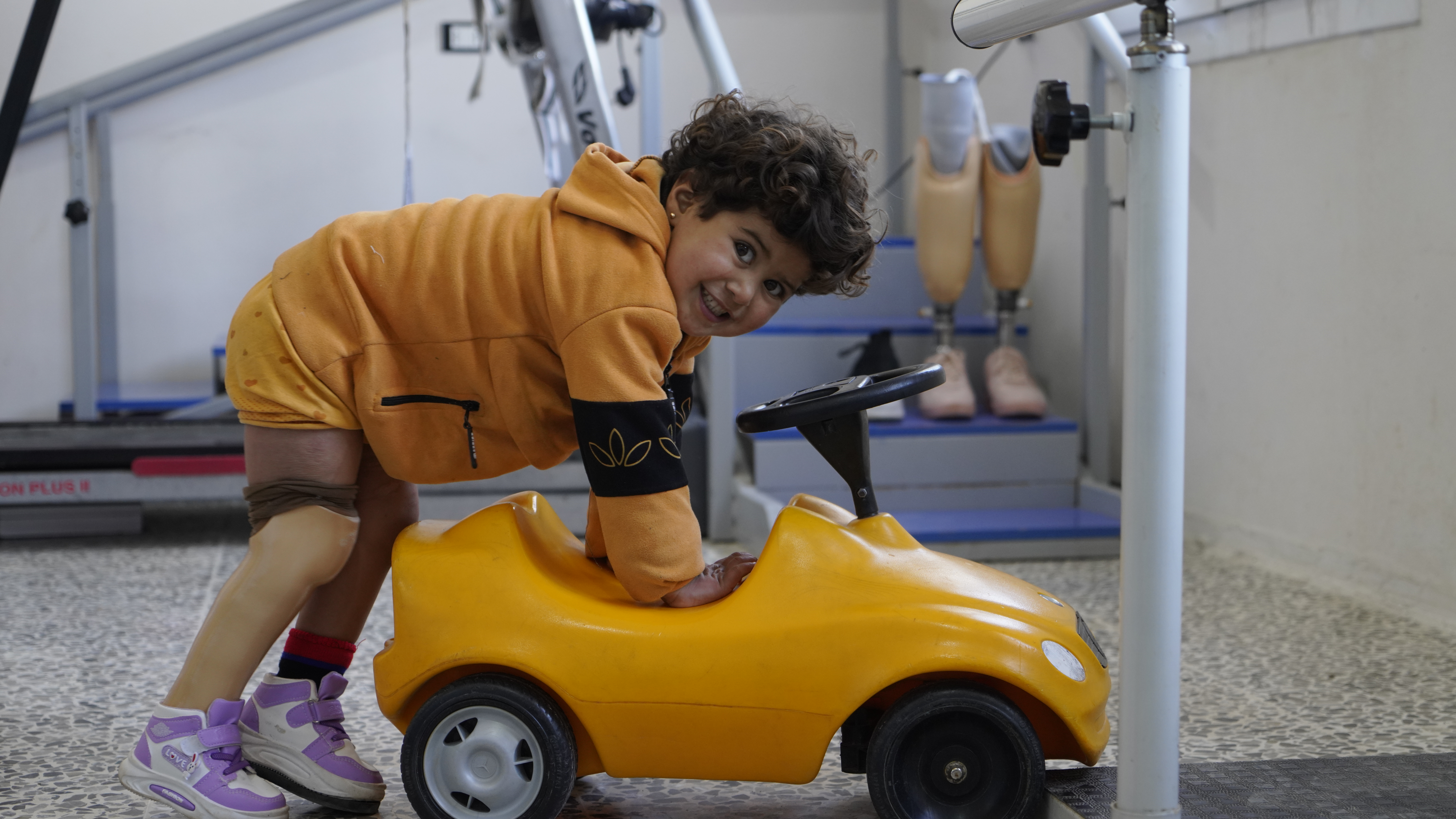 Noor during a rehabilitation session. She jumps on a little yellow car smiling at the camera at Aqrabat Hospital, HI’s partner in Northwest Syria. 