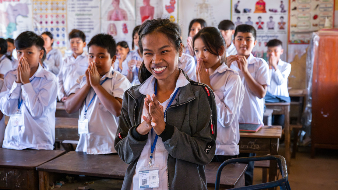 Close-up photo of a pupil smiling and waving with her hands clasped together. Behind her, other pupils can be seen in a classroom.