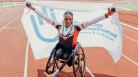 Young woman with no legs, smiling, sitting in a wheelchair on a track circuit, holding behind her a Humanity & Inclusion flag