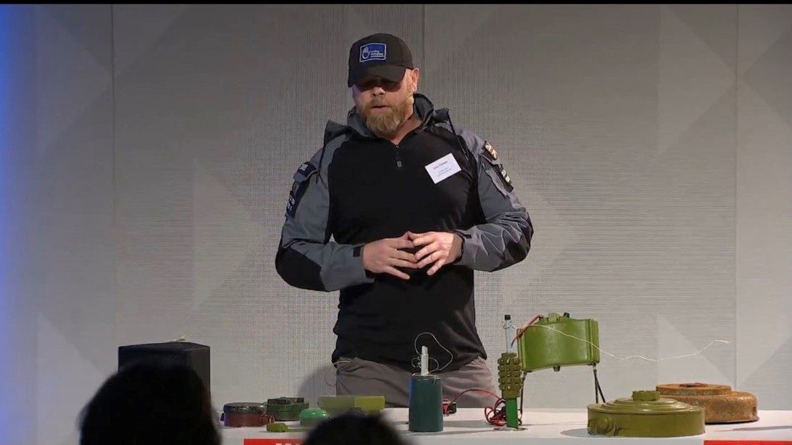 Man with a beard, wearing HI hat, and jacket, stands behind a table with different devices. 