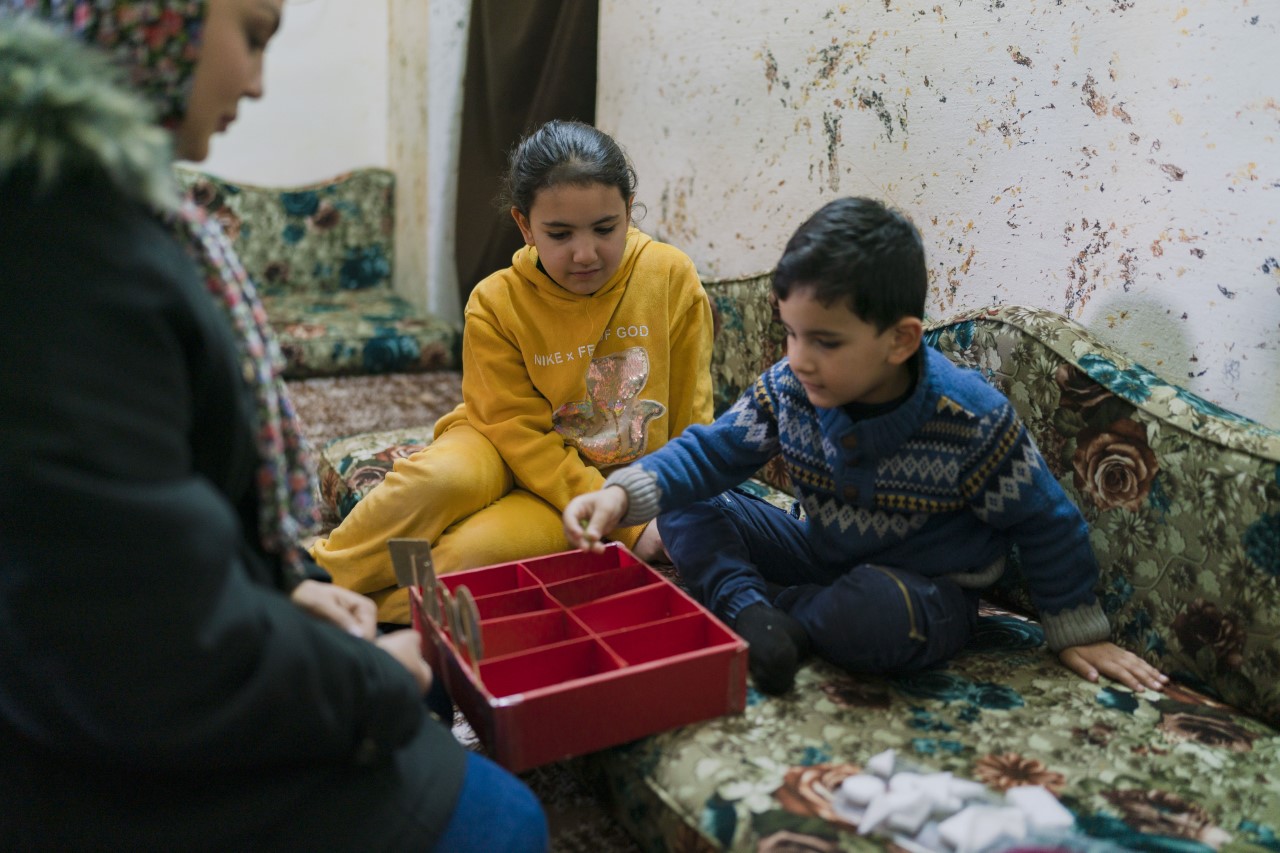 young boy wearing blue sweater placing an item in a red segmented box. he sits on a couch next to his sister, wearing a yellow hoodie, who watches on. they are opposite a woman wearing a head covering.