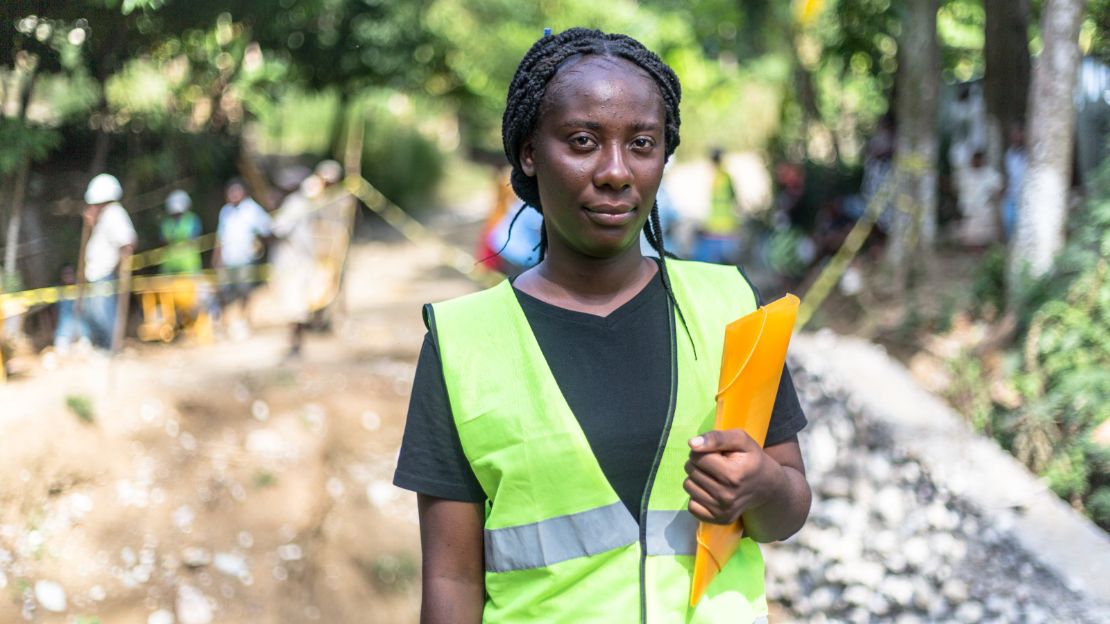 A black woman wearing a reflective vest holds a folder in front of a work site.