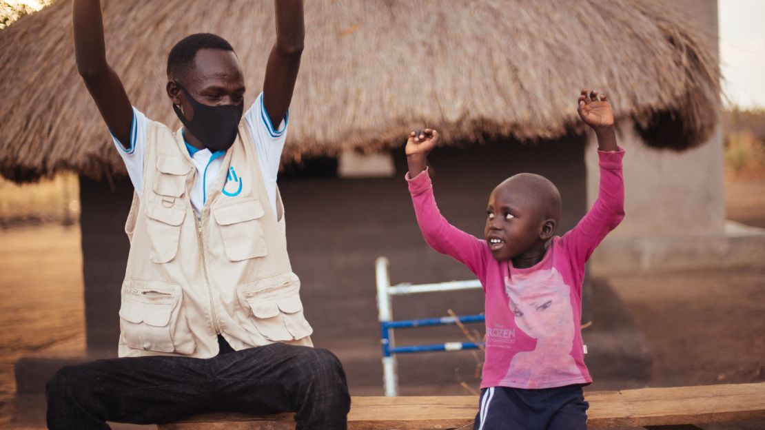 A male physician sits on a wooden bench with a little boy. They both smile as they hold their arms above their heads in a rehabilitation session. 