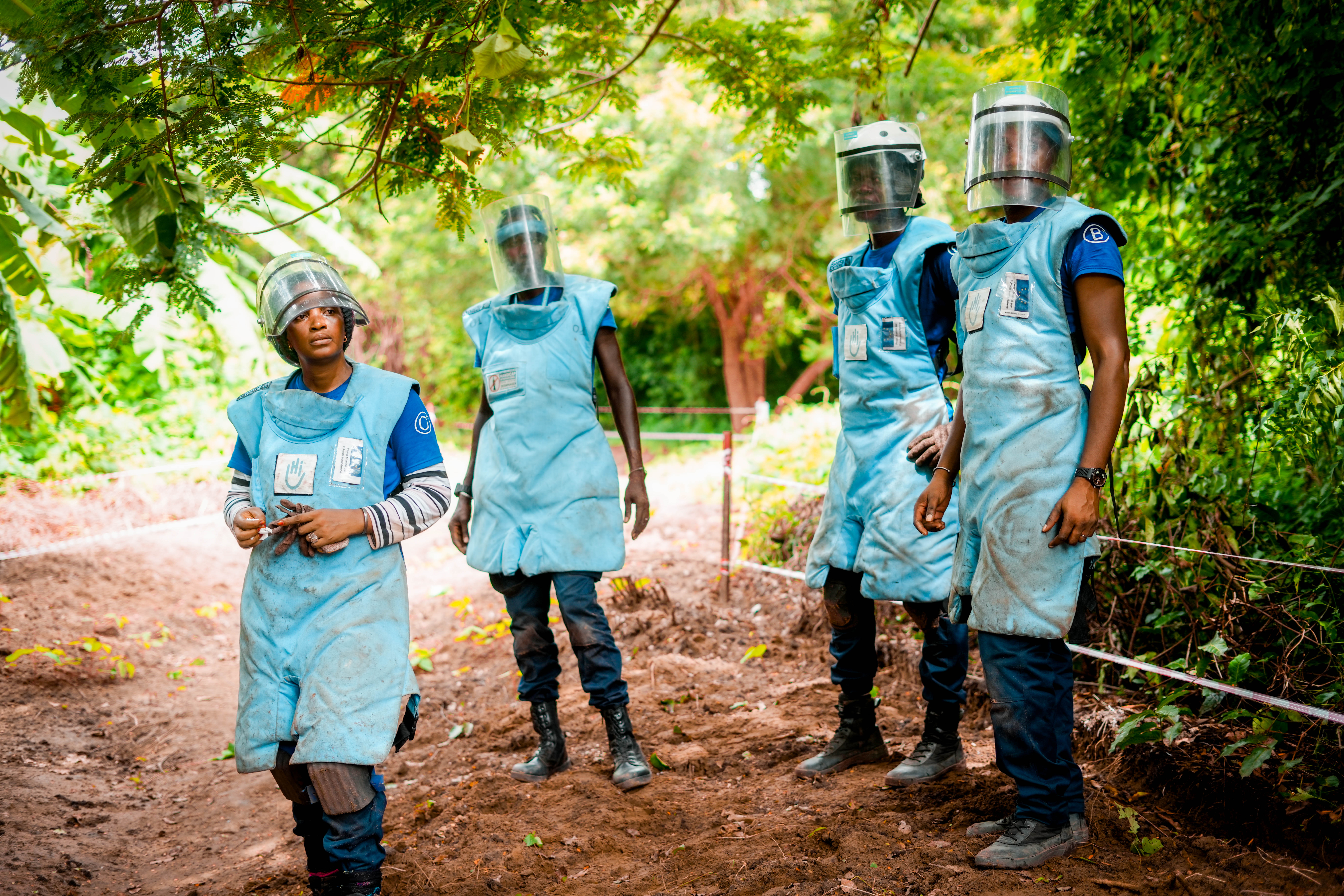 Four deminers in protective gear stand on a dirt track, surrounded by lush vegetation.