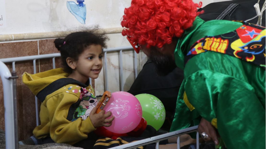 A little girl sits in a hospital bed playing with balloons during a visit from a clown