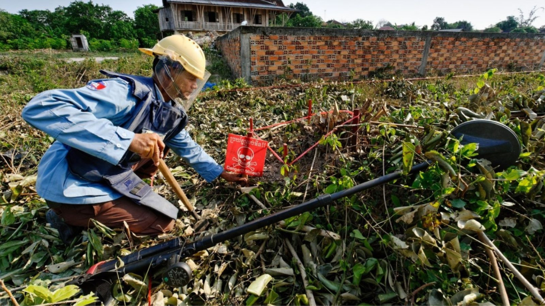 A mine clearance specialist wearing protective gear kneels on the ground in a Cambodian village placing a warning sign.