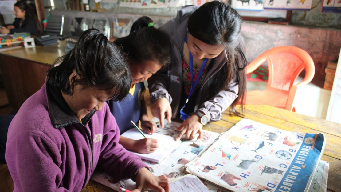 Two female students being guided in learning the English alphabet in a book by a specialist.