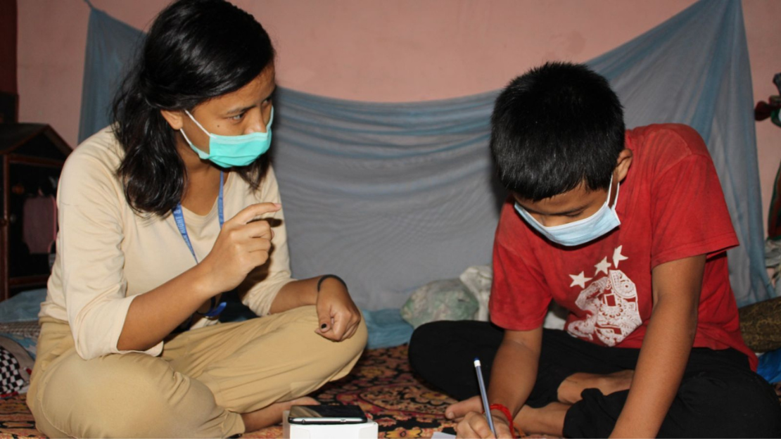 A woman and boy sit on the floor. She gestures to him in Nepali sign language as he writes on a notepad 