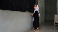 A young Afghan girl wearing a white head scarf, black outfit, and leg brace stands at a chalk board in a classroom.