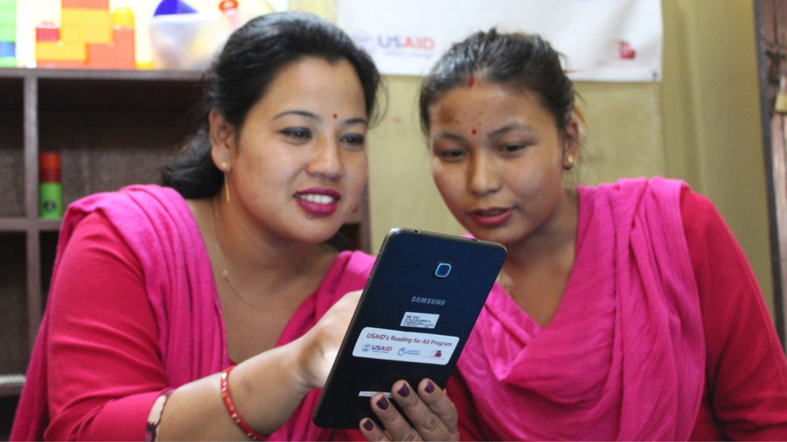 Two women both wearing bright pink sitting next to each other smiling using a tablet 