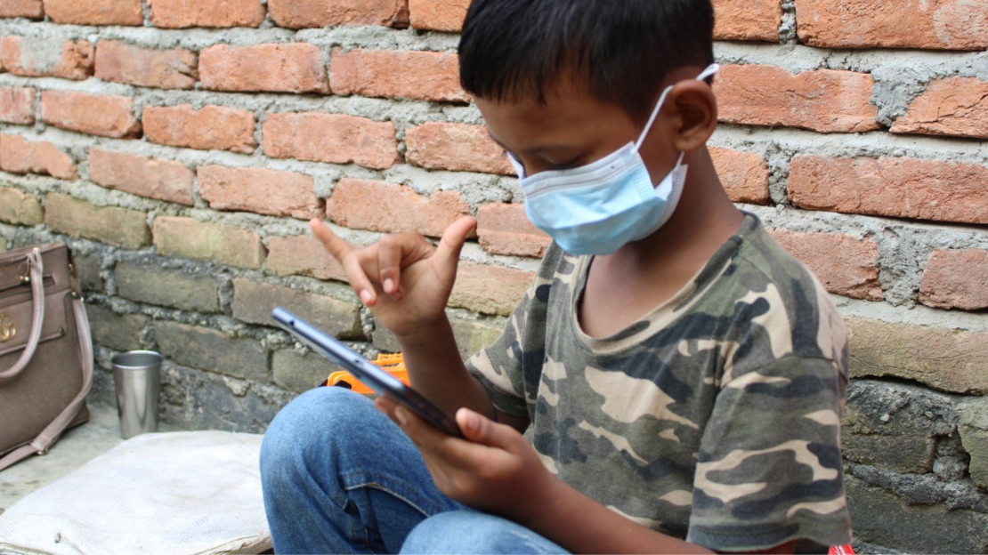 A young boy wearing a camo t-shirt and mask holding a phone in one hand and signing with his pinky and thumb with his other hand.