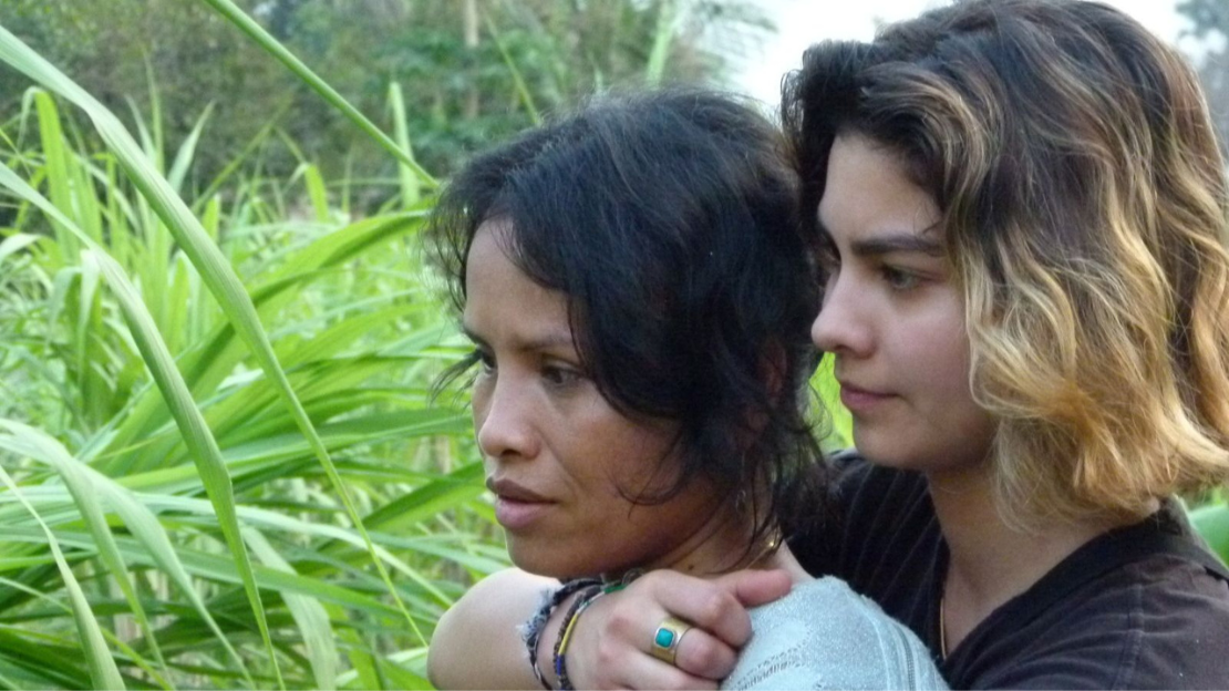 A young woman stands behind her mother wrapping her arm around her mother's chest.