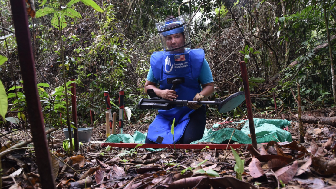 A woman deminer wearing a protective vest and face shield kneels on the ground in a wooded area in Colombia. She is holding a metal detector