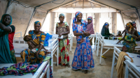 A group of women, all wearing colorful dresses and head coverings, stand in a medical tent. One woman holds her daughter in her arms. Another woman holds her baby.