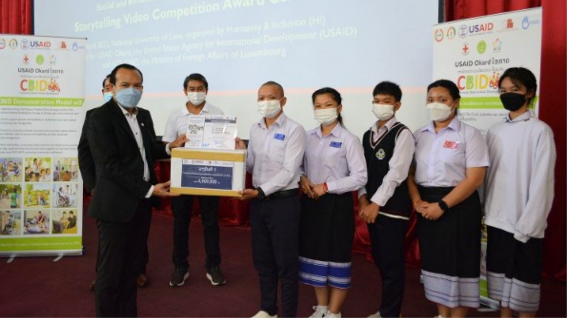 Group of winners accepting their award. Standing in front of two posters and a powerpoint screen between red curtains. Everyone wearing masks.