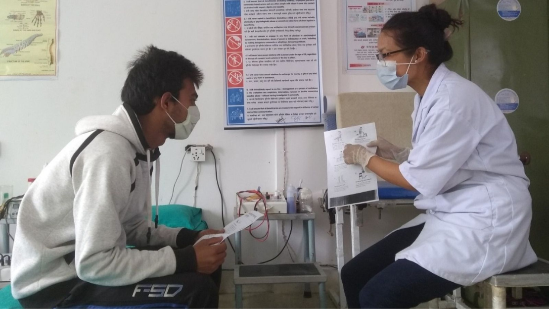 A female physical therapist wearing a white coat, glasses, and a mask provides instructions on different exercises to a rehabilitation patient wearing a grey sweatshirt and mask.