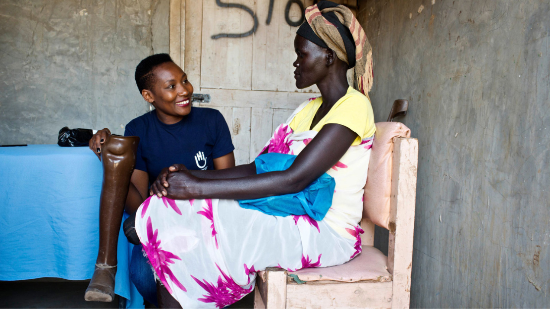 A woman, wearing a pink and white dress and a head covering, sits in a chair. Beside her is a smiling specialist wearing an HI tshirt holding a prosthetic leg.