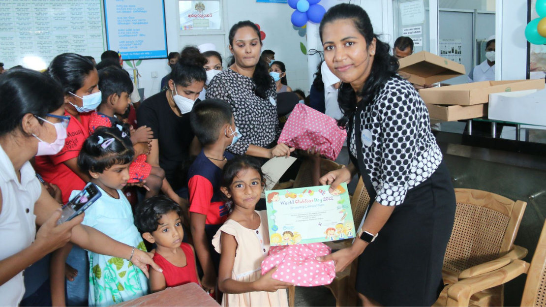 A woman smiles as she hands a world club foot day certificate to a smiling young girl at an event with other people in the background, some wearing masks. 