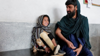 A young Afghan girl wearing a partial head covering and prosthetic leg smiles seated next to her father, a man wearing a black shawl with a black beard.