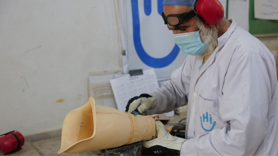 A man wearing a white lab coat, gloves, goggles on his forehead, a mask and ear protectors shapes part of an artificial leg.