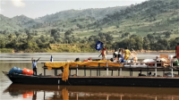 A group of men stand on a boat carrying humanitarian supplies along a waterway in the Central African Republic. One man is waving an HI flag. In the background are tree-lined hills.