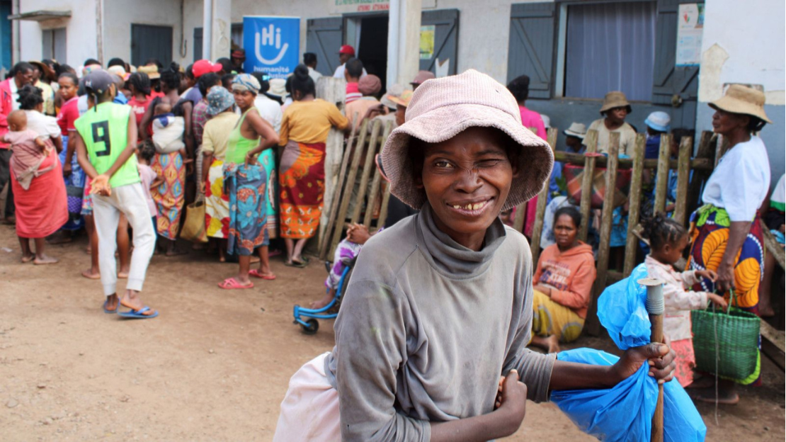 A woman wearing a hat stands in front of a crowd of people at a cash and other basic needs distribution. An HI poster is hung.