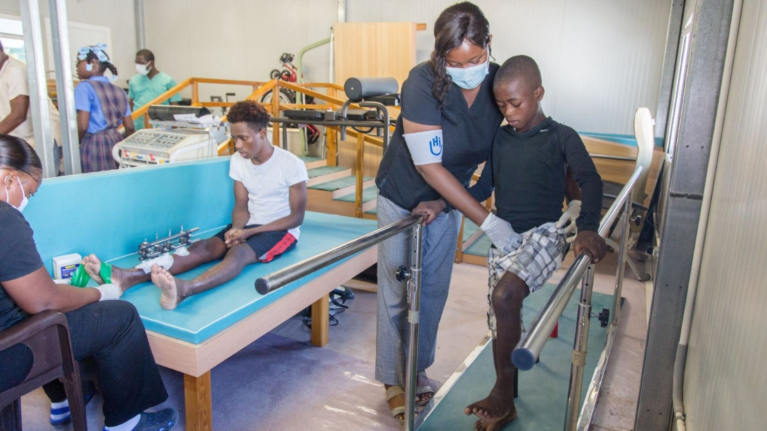 Woman wearing black scrubs, mask, and arm band with HI hand logo, assists a young boy on parallel bars. To the left, a teenage boy is on seated upright on a medical bench with a medical device on his leg and his foot being measured by a specialist. In the background are physical therapy devices, stairs, and a few other people facing away.