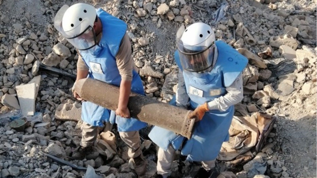 Two men wearing protective vests and face face shields carry a bomb amid rubble.