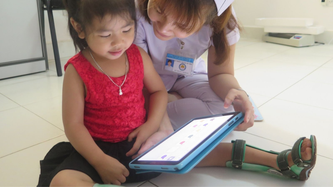 A young girl wearing a red top and a medical worker wearing lilac scrubs sit on the floor looking at a tablet 