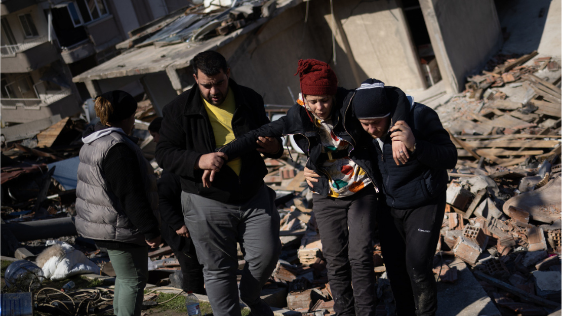 Two men supporting an injured woman walking in front of a collapsed building