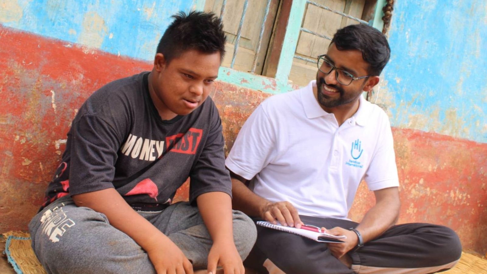 Two young men sitting in front of a building. The one on the left wearing an HI polo and glasses and holding a note pad smiling, The one on he left wearing a graphic t-shirt. 