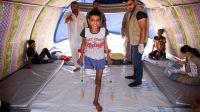 Abdel Rahman stands with crutches in his tent, while HI physical therapist Mohammed looks on.
