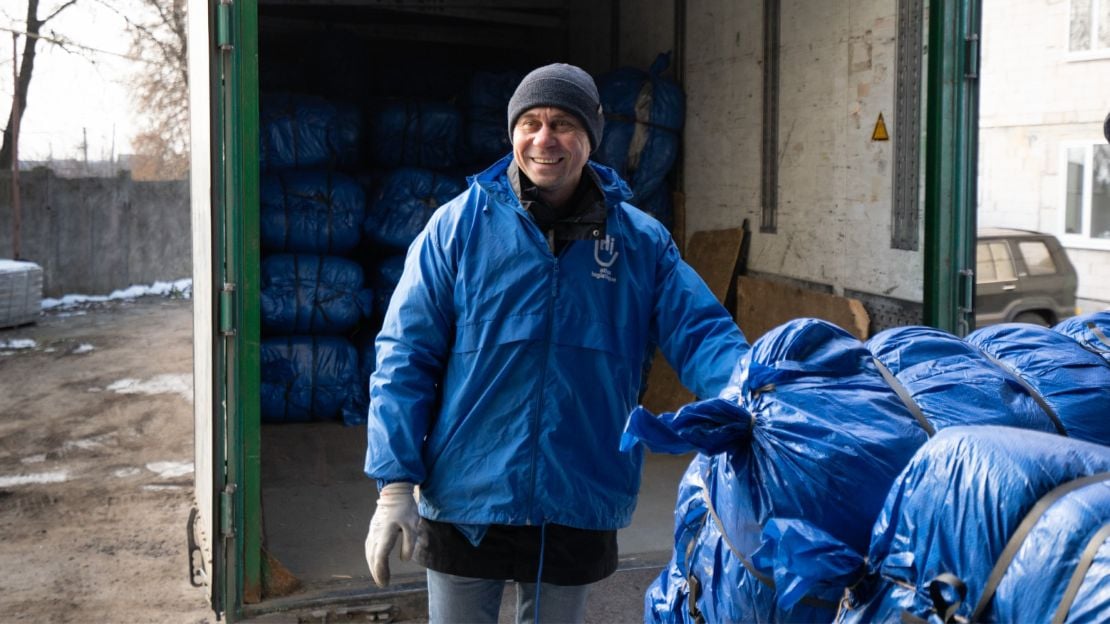 A man wearing a blue HI jacket and gloves smiles. He's standing in front of a box truck and next to a pile of blue bags containing humanitarian supplies.